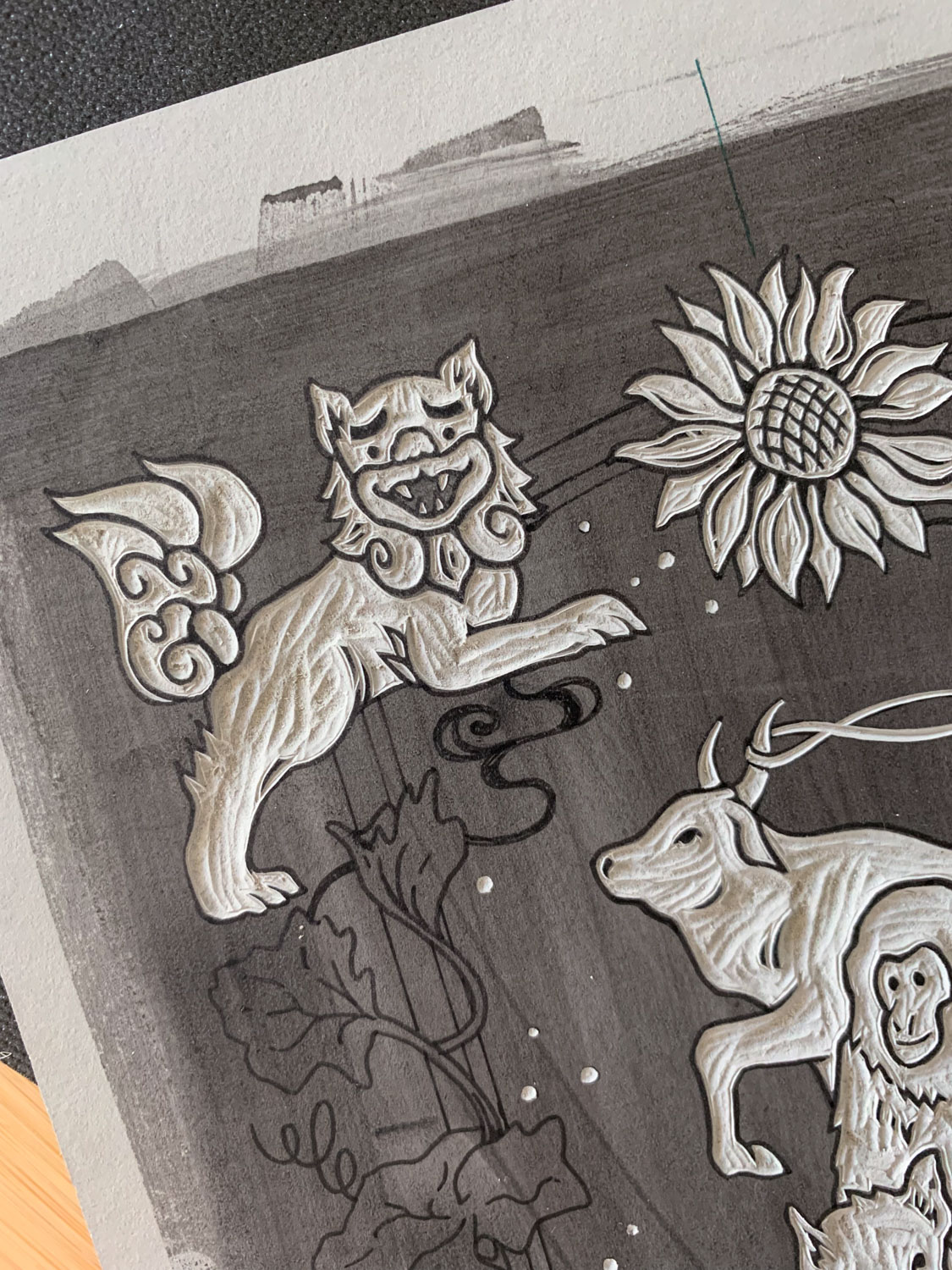 A closeup view of the partially carved grey linoleum block showing a carved shisa, sunflower, zodiac ox, and partial monkey face. The rest of the block is stained a darker grey and has pen drawings outlining the rest of the design.