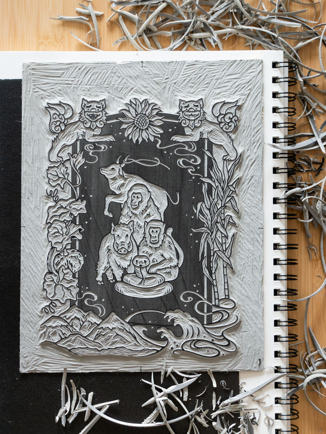 A head-on view of the final carved linoleum block. The block sits on top of a black non-slip pad and a large white sketchbook, which are surrounded by lots of linoleum shavings.