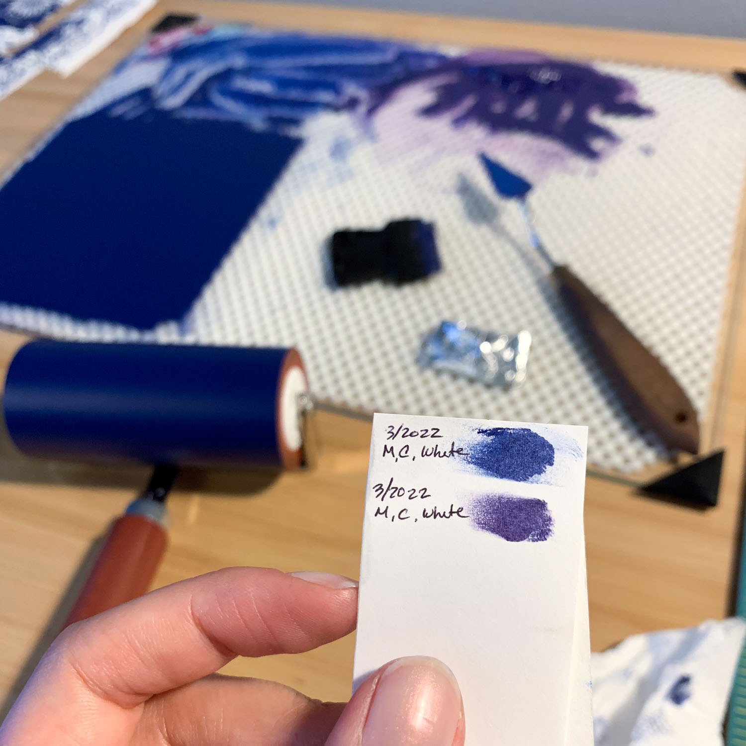 A hand holds a white piece of paper with two smears of paint, one blue and one purple. Both are labeled '3/2022 M, C, White'. In the background there is a glass inking plate with swatches of blue and purple ink, as well as a brayer covered in blue ink.