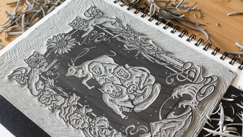 A grey linoleum block sitting on top of a notebook. The linoleum is carved with zodiac animals and foliage.