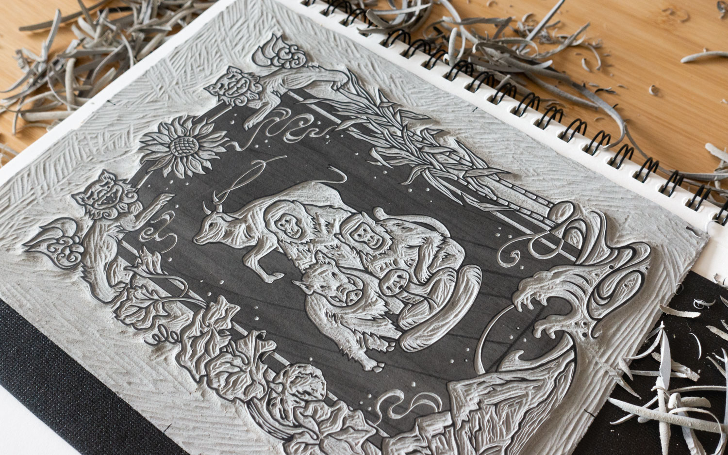 A grey linoleum block sitting on top of a notebook. The linoleum is carved with zodiac animals and foliage.