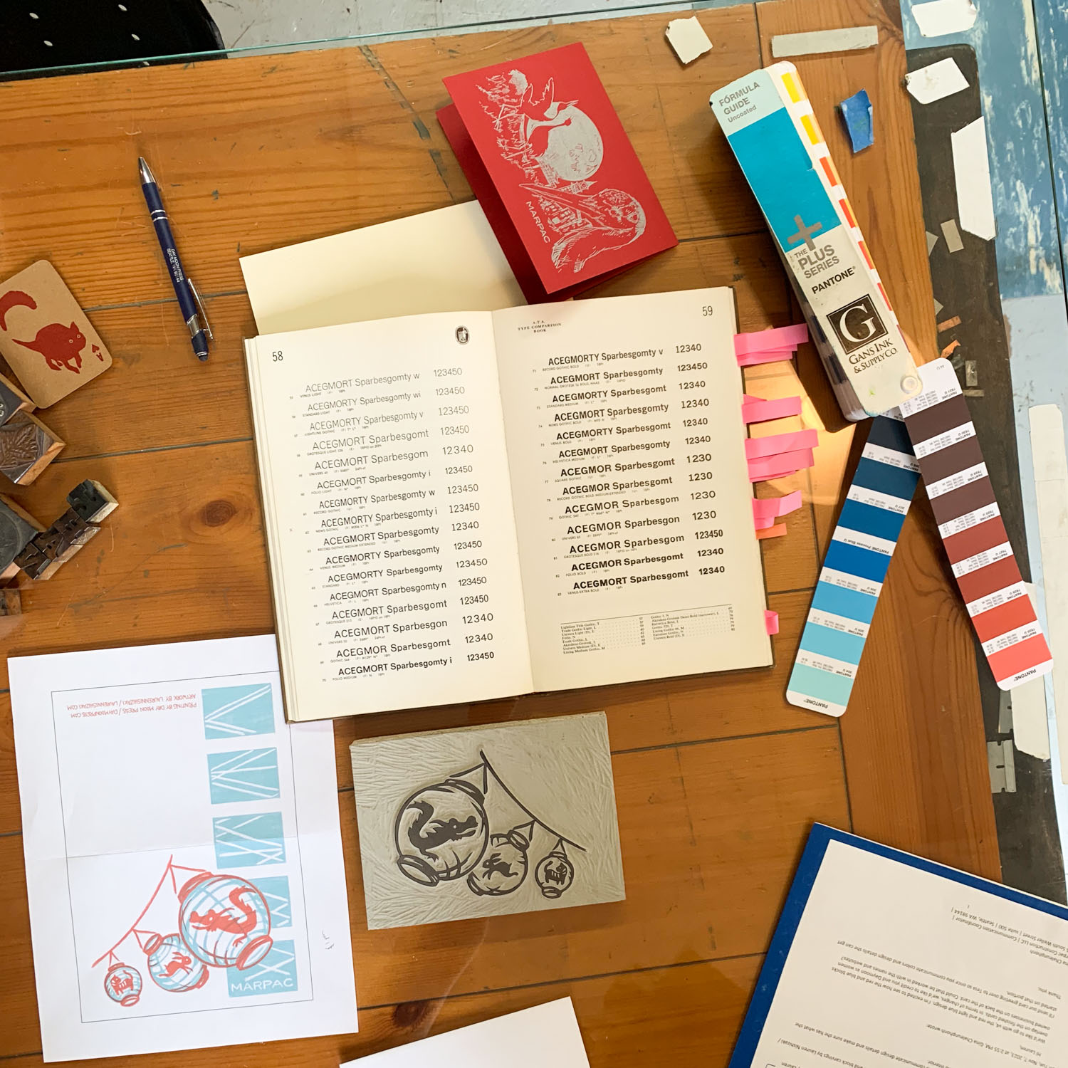 A book with examples of type of different fonts and sizes, surrounded by a Panton swatch book open to red and blue, surrounded by a printout of the card design, a carved linoleum block, and various other papers.