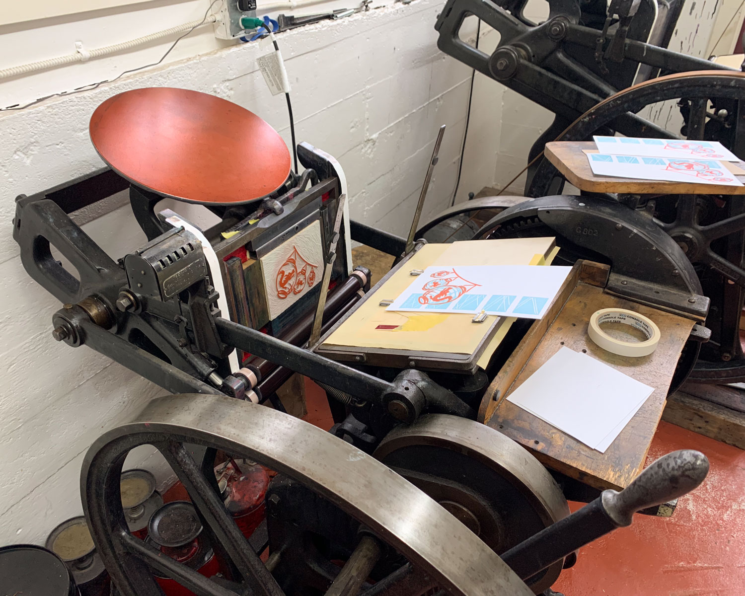 A platen letterpress machine; the ink plate is covered in red ink and a linoleum block is mounted in the press, also covered in red ink.