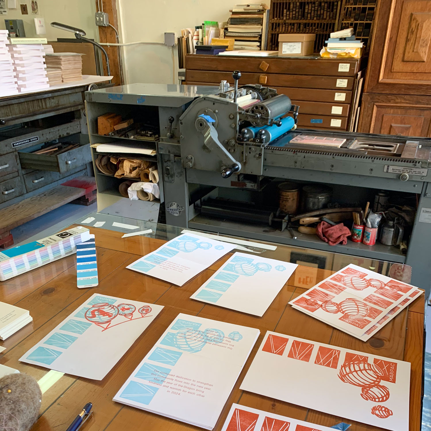 Several rectangular sheets of white paper printed with different test variations of the card design. A large cylinder press is visible in the background, surrounded by piles of paper and cases of type.