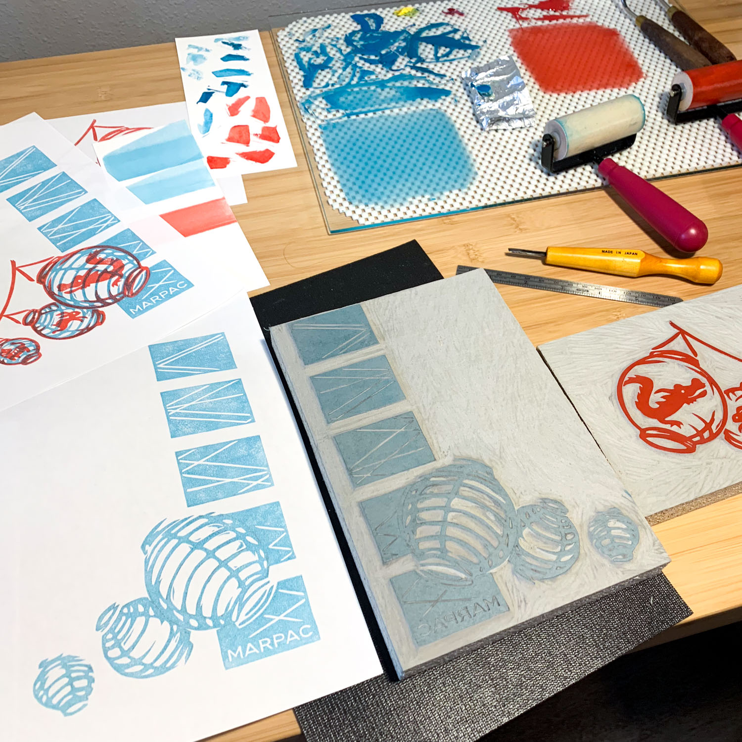 A desk covered in carved linoleum blocks, glass inking plate (with red and light blue inks rolled out), brayers, and test prints on rectangular sheets of white paper.