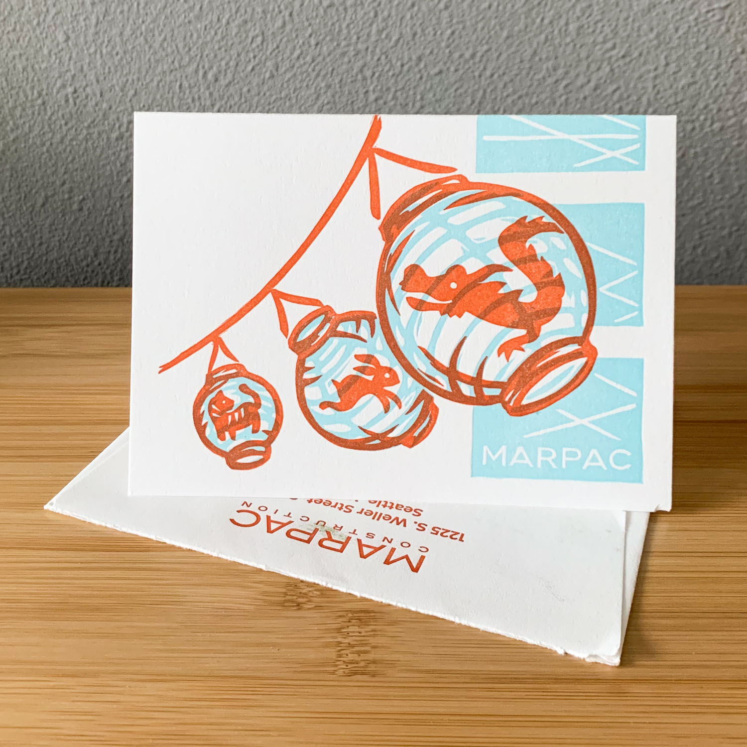 A horizontal card propped open on top of a white envelope with Marpac's return address in red. The card is white with a red and blue illustration of 3 lanterns (depicting a tiger, rabbit, and dragon) against a geometric background. The card and envelope sit on a wood tabletop.