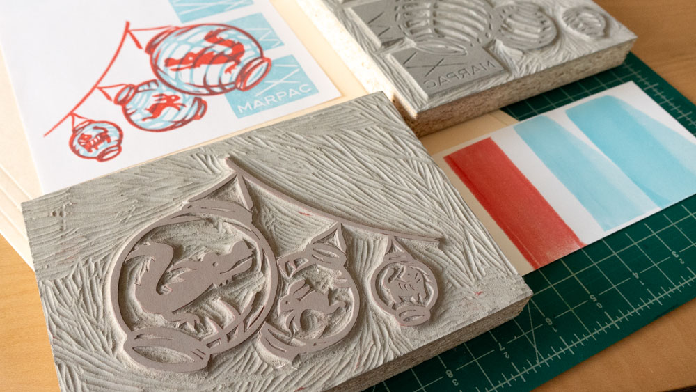 Two mounted and carved linoleum blocks laid out with two sheets of white paper printed in red and blue. The blocks are carved with different patterns that when combined produce the printed image on the top left sheet of paper.