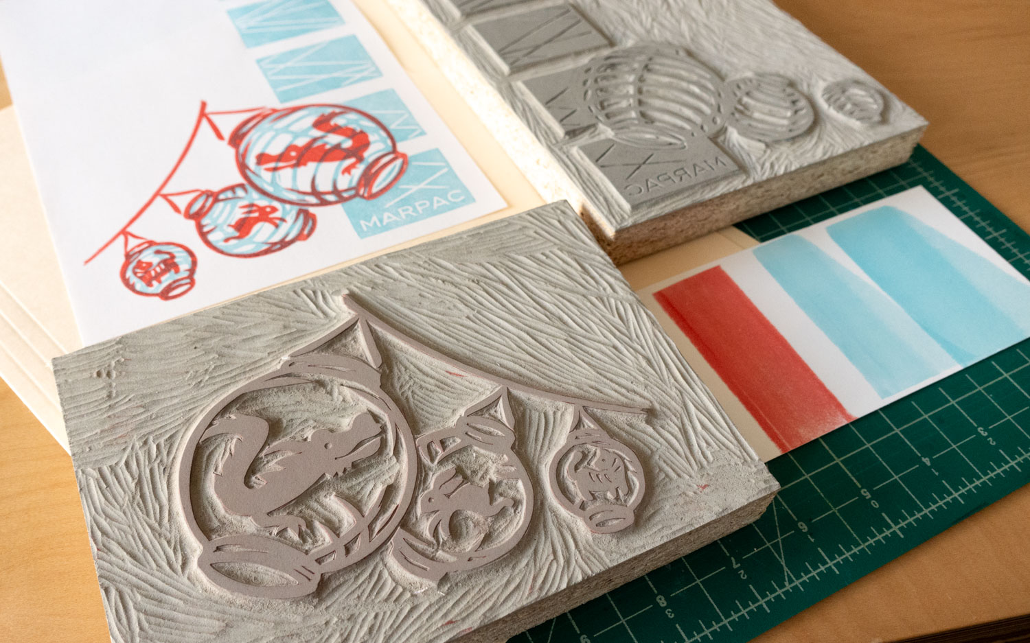 Two mounted and carved linoleum blocks laid out with two sheets of white paper printed in red and blue. The blocks are carved with different patterns that when combined produce the printed image on the top left sheet of paper.