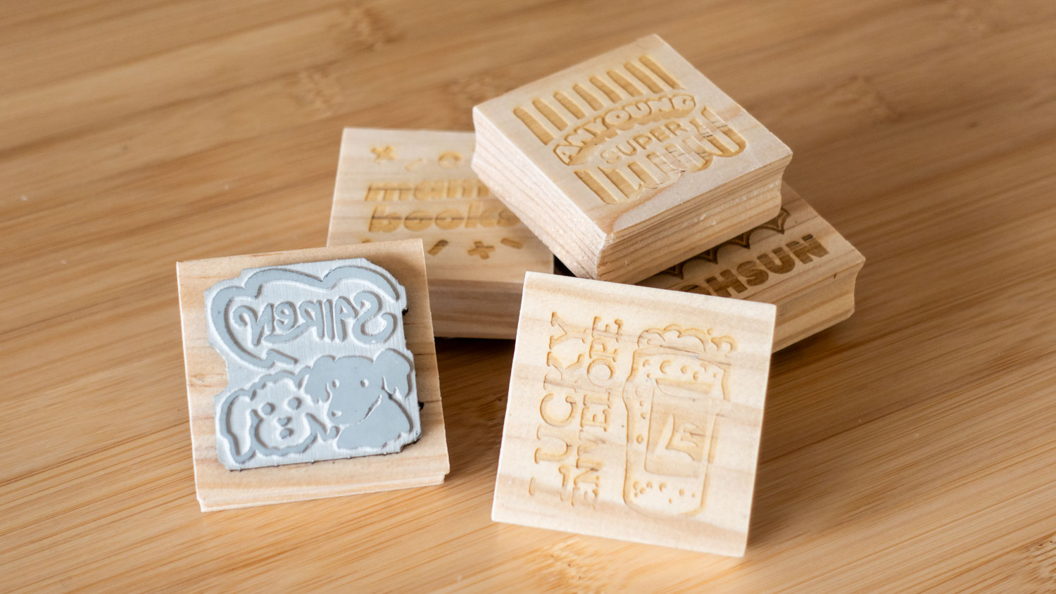 A jumble of square stamps, with grey rubber stamps and laser-engraved wood backing. The visible stamps read 'Sairen' (in reverse), 'Lucky Envelope', and 'Mam's Books'.