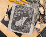 A messy desk covered with a carved linoleum block of a boar and maple tree. The block is surrounded with carving tools and linoleum shavings.