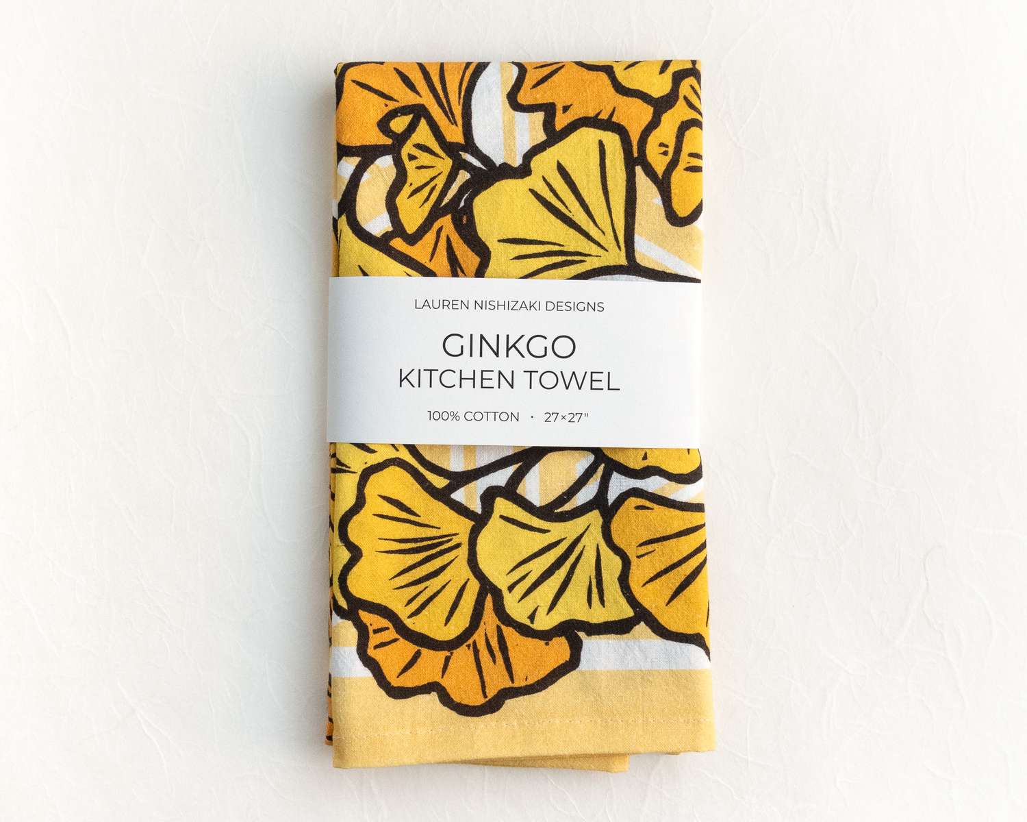 A folded kitchen towel featuring a yellow and black illustration of ginkgo leaves. A white belly band reads 'Lauren Nishizaki Designs; Ginkgo kitchen towel; 100% cotton; 27x27