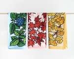 Three colorful kitchen towels hanging on a wooden dowel. From left to right, they depict blue and green hydrangea, red Japanese maple, and yellow ginkgo