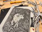 A messy desk covered with a partially carved ox blockprint, carving tools, and grey linoleum shavings.