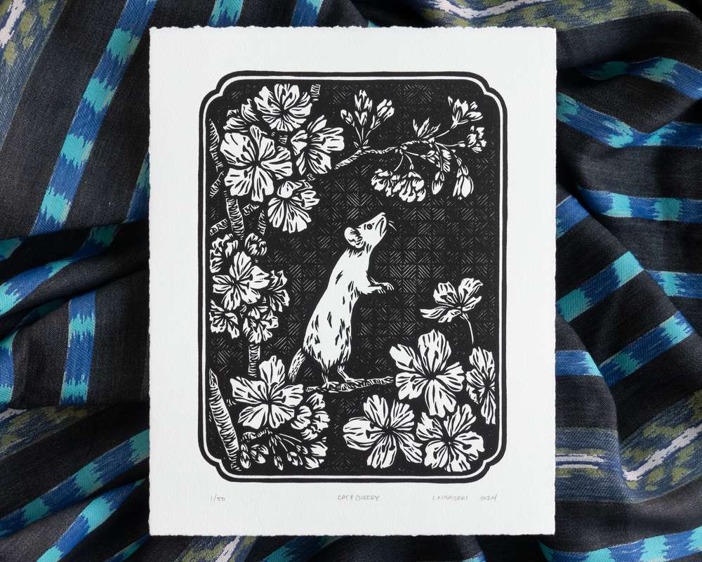 Black and white illustration of a rat standing in a cherry tree gazing at cherry blossoms. The white paper sits on top of rumpled blue striped fabric.