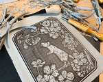 A messy desk covered with a partially carved rat blockprint, carving tools, and grey linoleum shavings.