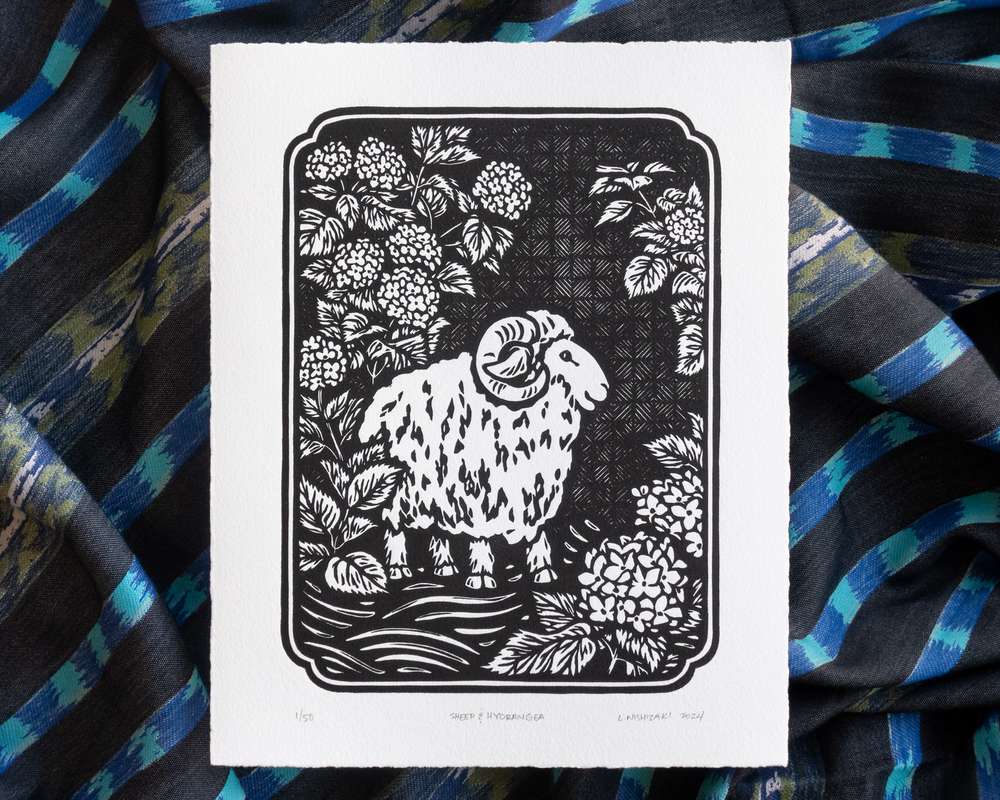 Black and white illustration of a sheep amogst hydrangea bushes. The white paper sits on top of rumpled blue striped fabric.