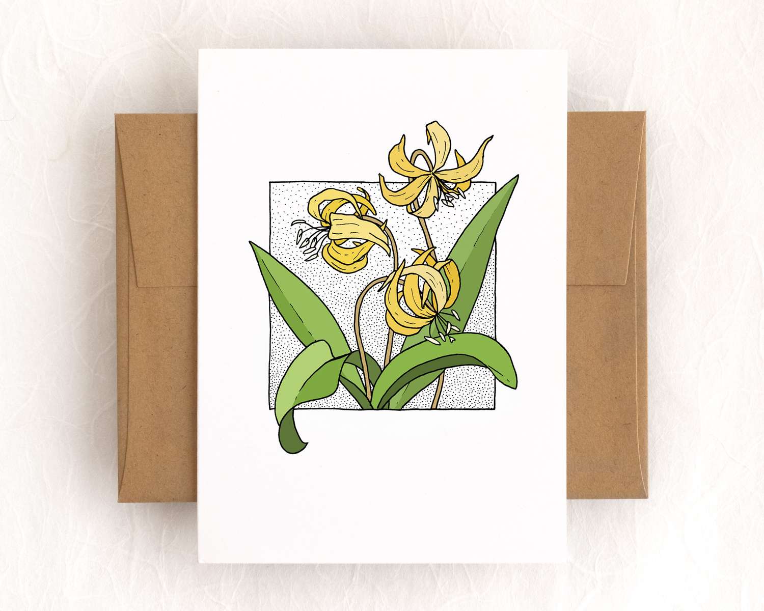 A vertical white card depicts a spray of yellow flowers surrounded by long spade-like leaves. The card sits on top of a brown envelope, which lies on top of a white backdrop.