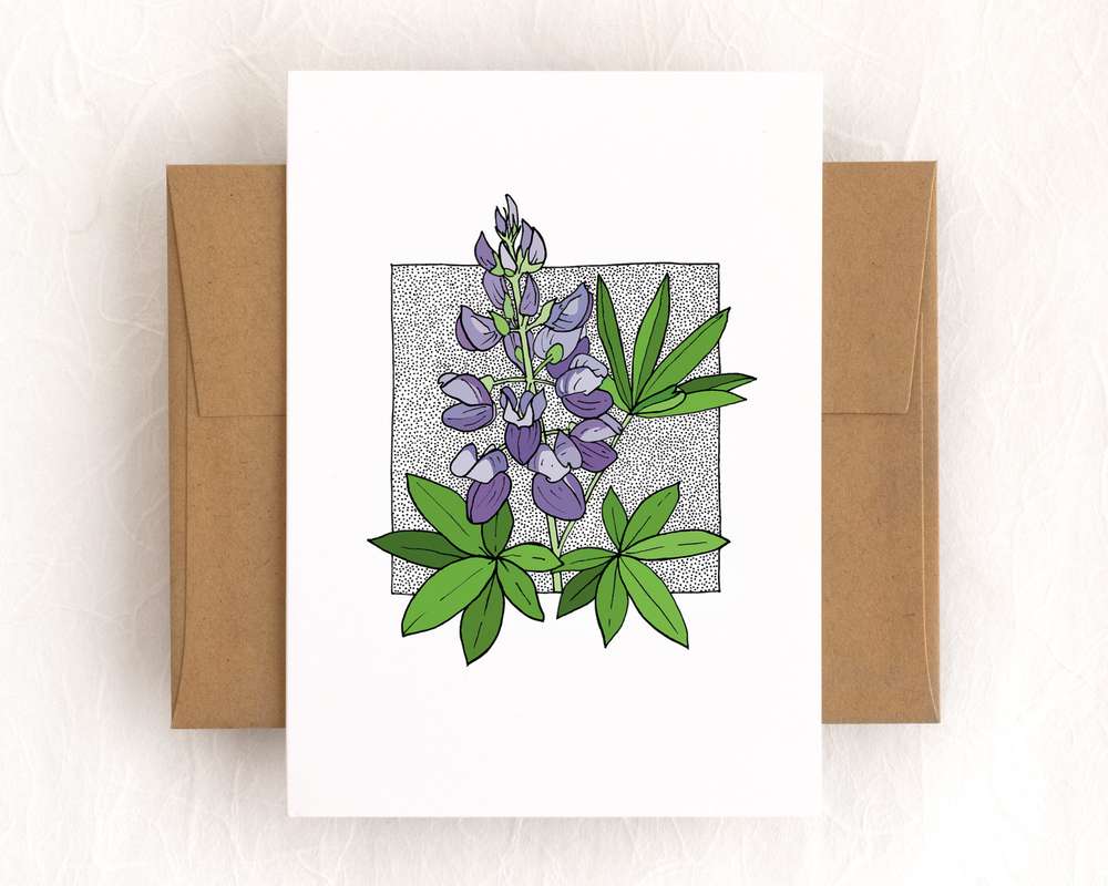 A vertical white card depicts a spray of lupine flowers surrounded by lupine leaves. The card sits on top of a brown envelope, which lies on top of a white backdrop.