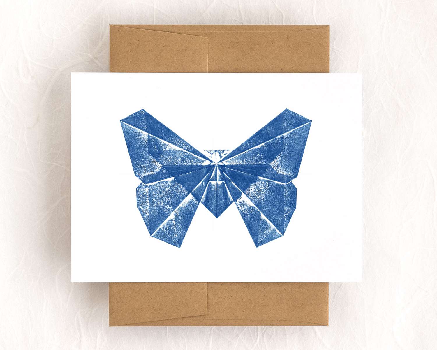 A horizontal white card depicts a bright blue butterfly, rendered in blocky and patchy triangles. The card sits on top of a brown envelope, which lies on a white backdrop.