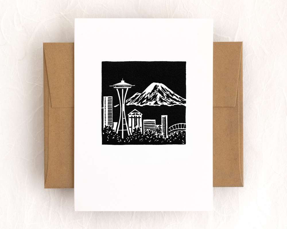 A vertical white card with a black and white depiction of the Seattle skyline and Mt Rainier rising in the distance. The card sits on top of a brown envelope, which lies on top of a white backdrop.