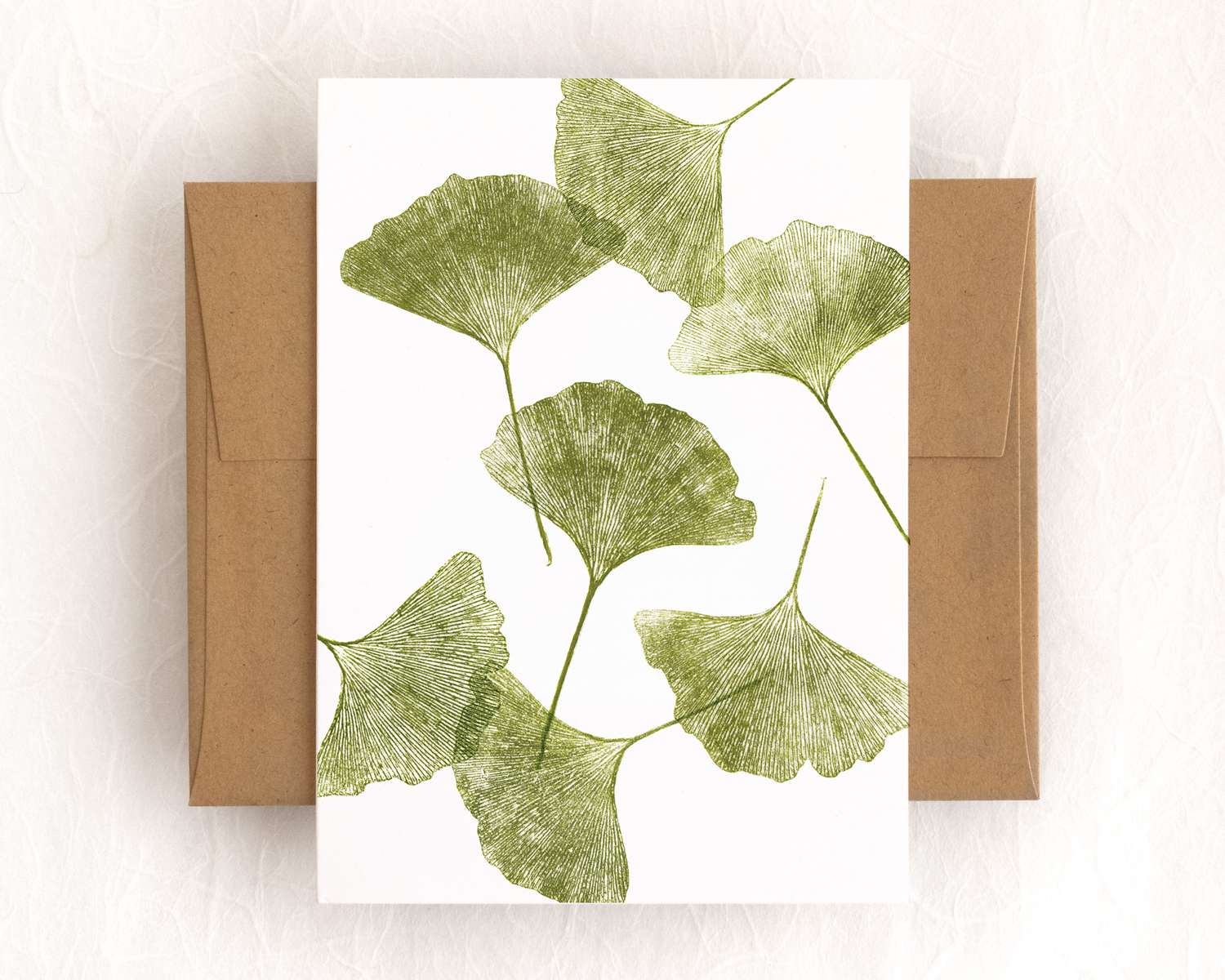 Seven green ginkgo leaves are scattered across the surface of a vertical white greeting card. The card sits on top of a brown envelope, which lies on a white backdrop.