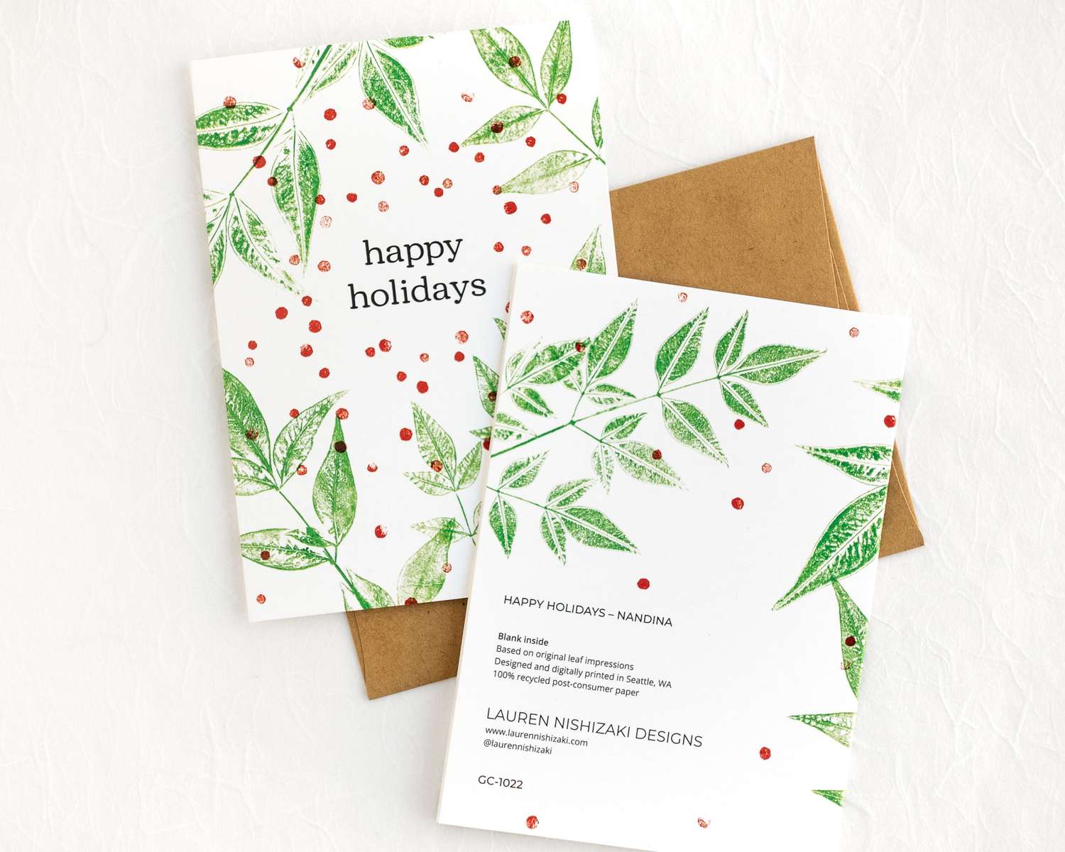 Two cards sitting on a brown envelope. One card shows the front, with green leaves and text that reads 'happy holidays'; the other shows the back with various text.