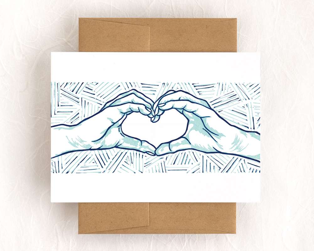 A horizontal white card depicts a blockprinted pair of hands making a heart shape. The design is done in navy and light blue. The card sits on a brown envelope, which lies on a white backdrop.