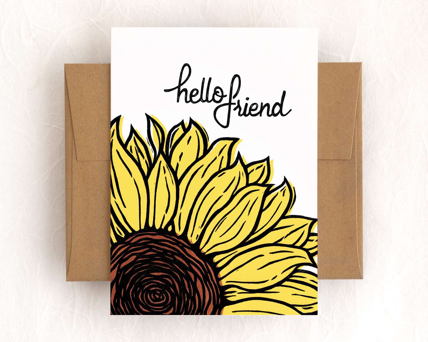 A vertical white card depicts a blockprinted yellow and black sunflower below text that reads 'Hello Friend'. The card sits on top of a brown envelope, which lies on top of a white backdrop.