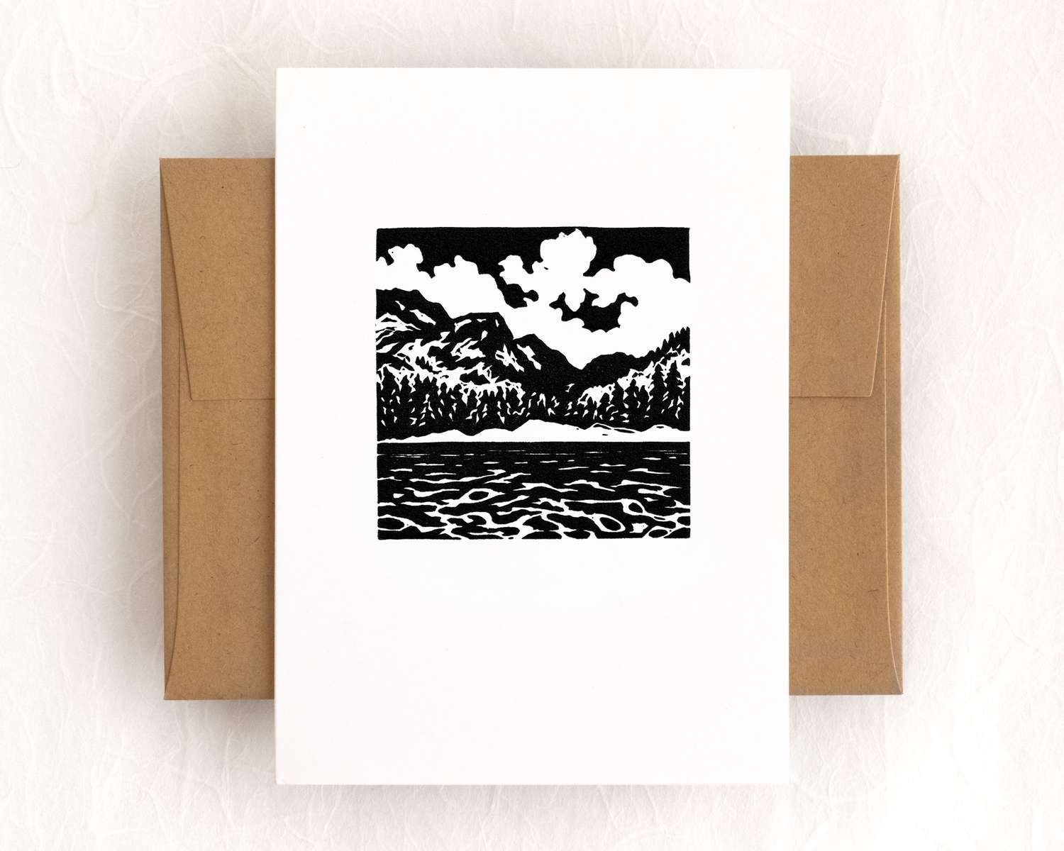 A vertical white card with a black and white depiction of a mountain lake. The card sits on top of a brown envelope, which lies on top of a white backdrop.