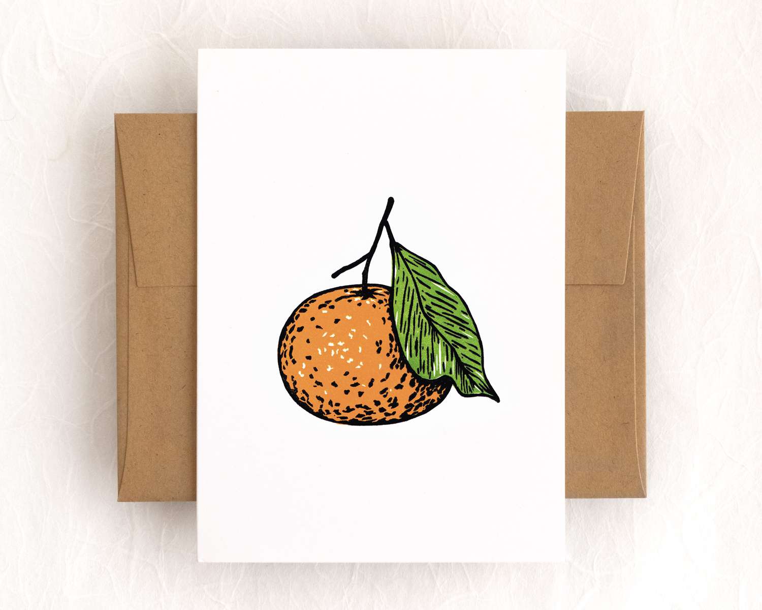 A vertical white card depicts a mandarin orange using bold black lines filled in with orange and green. The card sits on top of a white envelope, which lies on a brown backdrop.