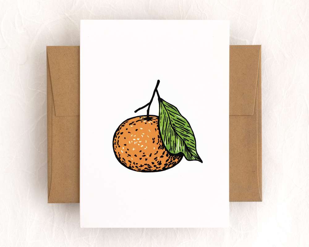 A vertical white card depicts a mandarin orange using bold black lines filled in with orange and green. The card sits on top of a brown envelope, which lies on a white backdrop.