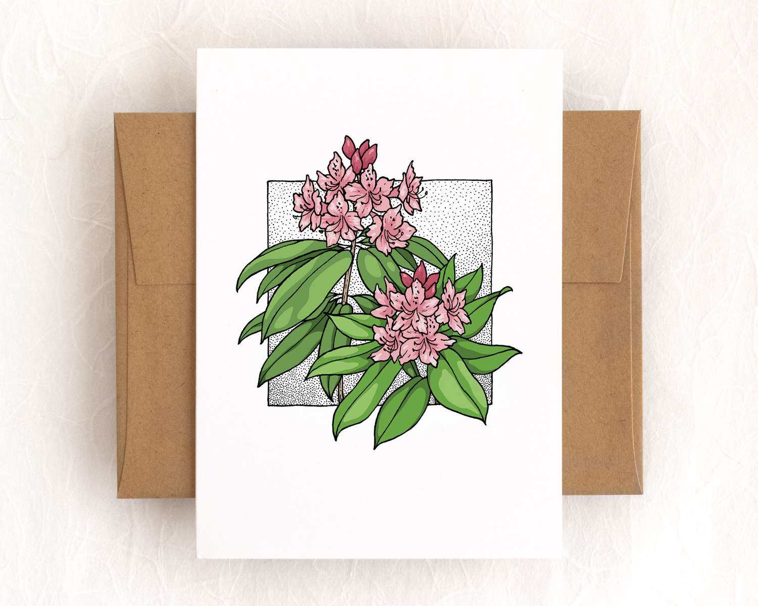 A vertical white card depicts two pink rhododendron flower clusters with green leaves. The card sits on top of a brown envelope, which lies on top of a white backdrop.