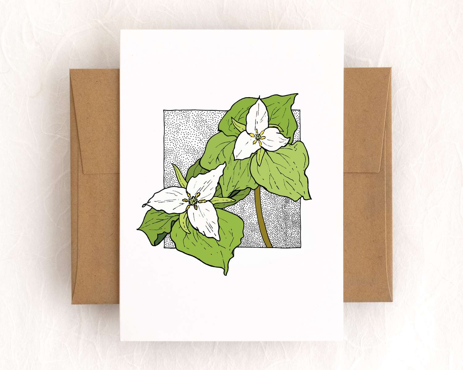 A vertical white card depicts two trillium flowers against a black stipple background. The card lies on top of a brown envelope, which lies on top of a white backdrop.