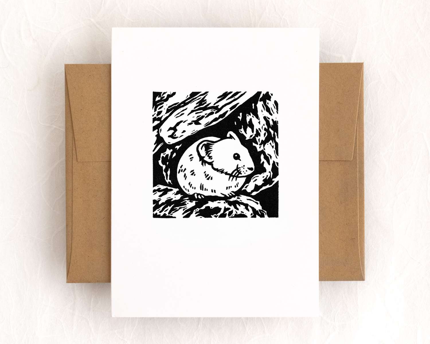 A vertical white card with a black and white pika sitting against rocks. The card sits on top of a brown envelope, which lies on top of a white backdrop.