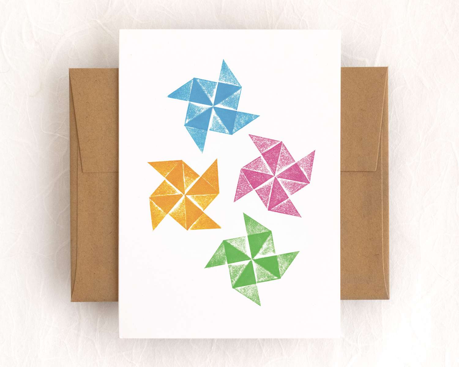 Four origami pinwheels – colored blue, orange, magenta, and green – are scattered across the surface of a white vertical card. The card lies on top of a brown envelope, which lies on a white backdrop.