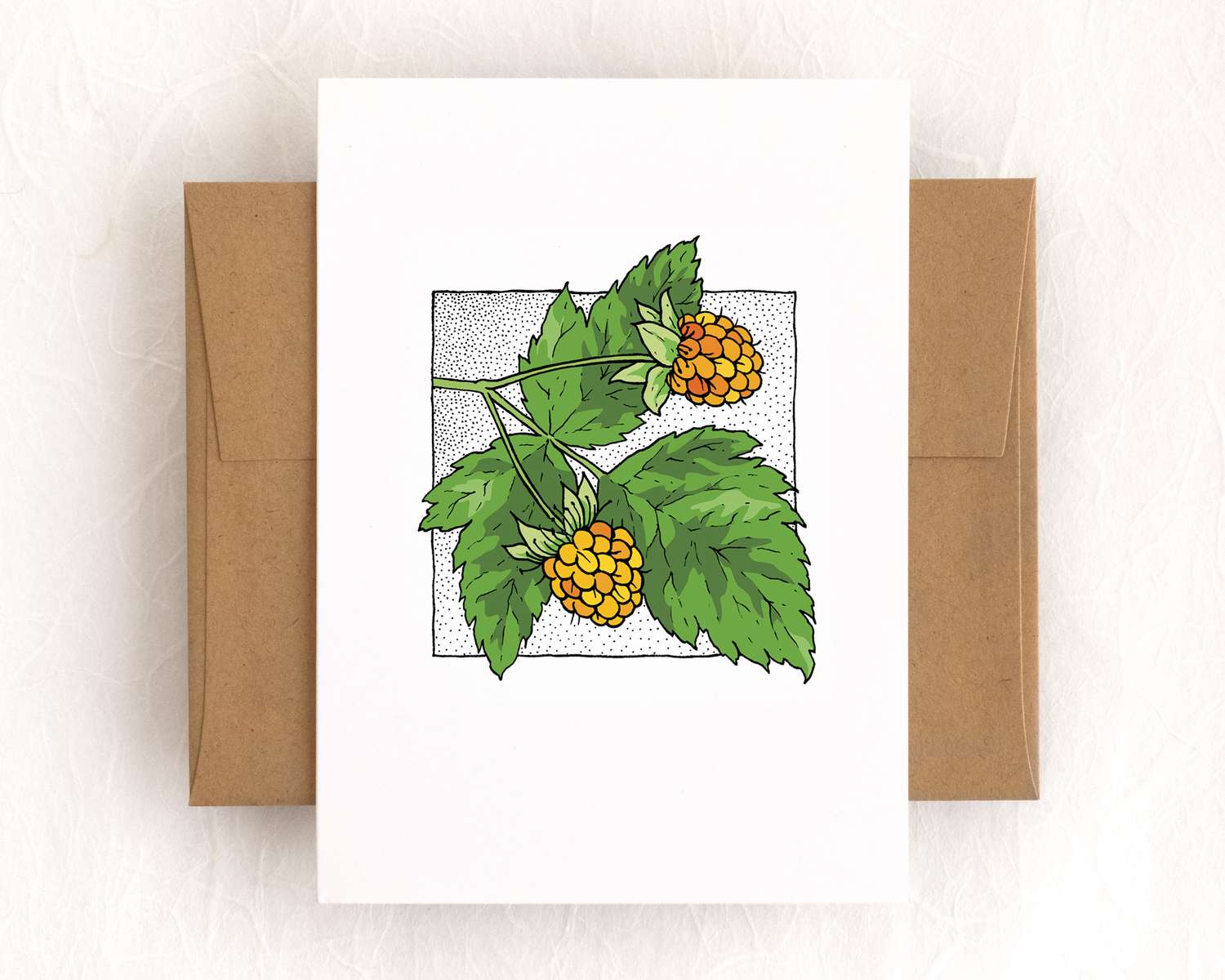 A vertical white card depicts two yellow-orange berries and green leaves. The card sits on top of a brown envelope, which lies on top of a white backdrop.