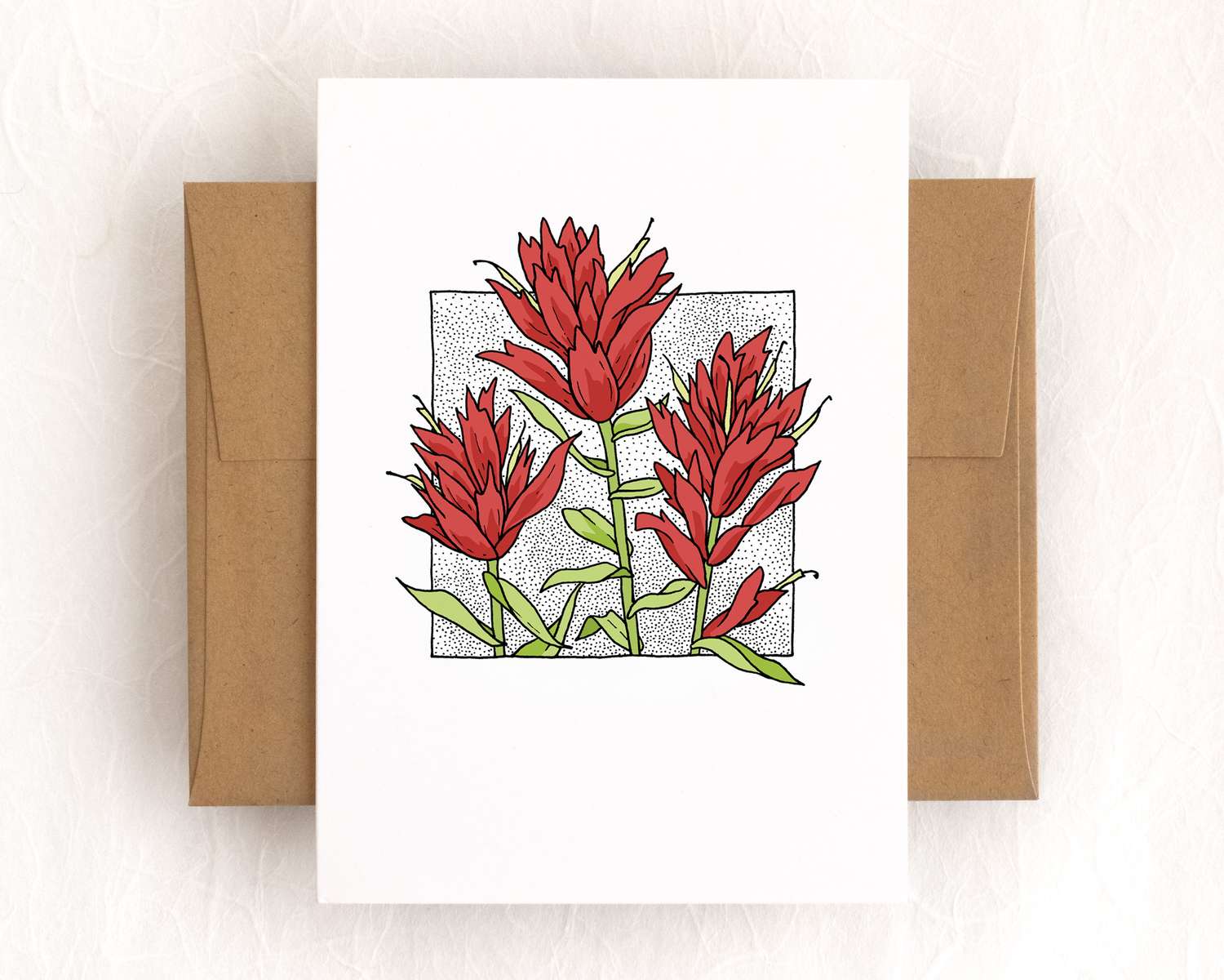 A vertical white card depicts three tall red flowers. The card sits on top of a brown envelope, which lies on top of a white backdrop.