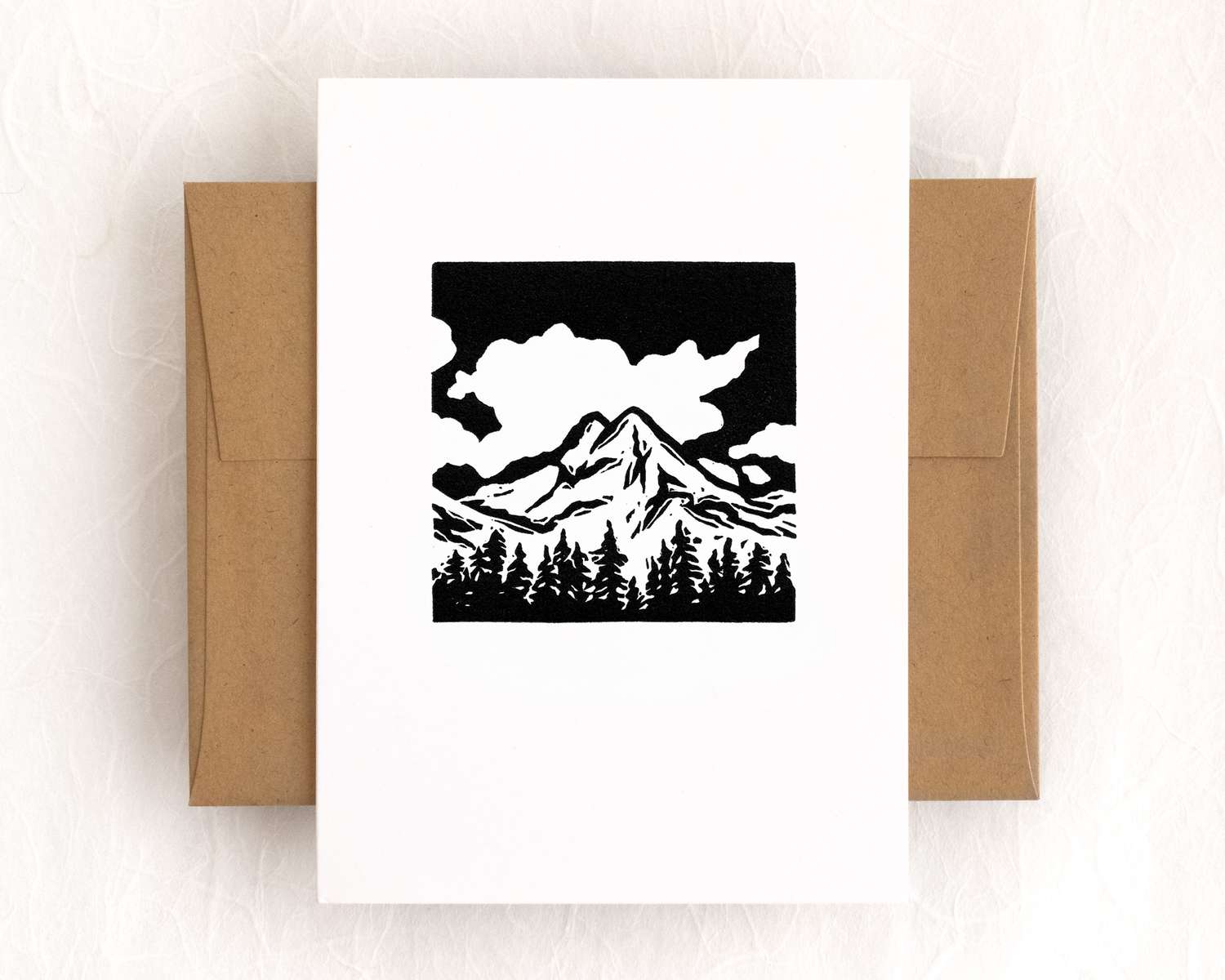 A vertical white card with a black and white depiction of a mountain peak and trees. The card sits on top of a brown envelope, which lies on top of a white backdrop.