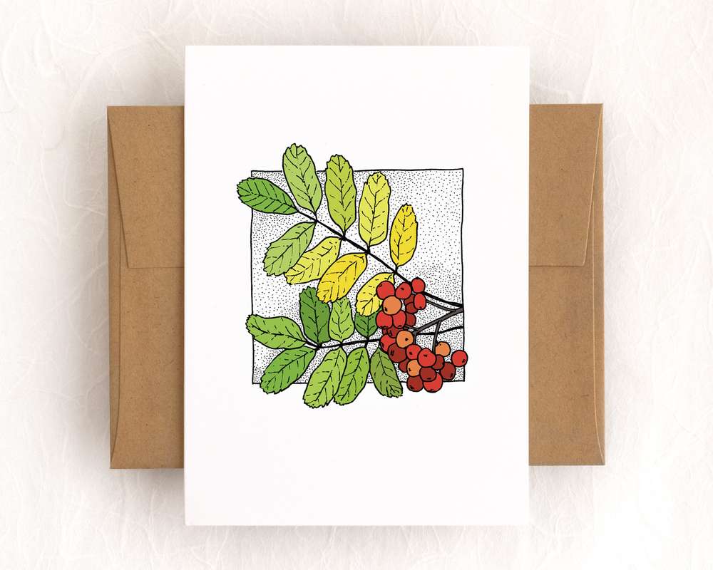 A white vertical greeting card, depicting two branches of green and yellow leaves and a spray of red berries. The card sits on top of a brown envelope, and both sit on a white backdrop.